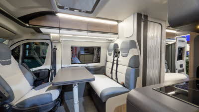 The Swift Carrera 244 has a grey floor and its upholstery is pale grey and blue. The cabinets are wood with white handles. The rising roof is open. Towards the rear there is lounge and one of its rear doors are open.