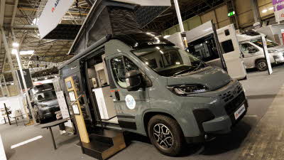 The Adria Twin Sports 600 SPB is grey, its rising roof is extended.  The sliding door is open and there are two steps to gain easy access.  There is an interactive stand.