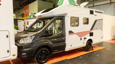 The Pilote Atlas A630G has a grey cab with a white coachbuilt body.  There are black, grey and red decals on its body.  Its skylights are open and there are two green and white striped umbrellas on its roof. 