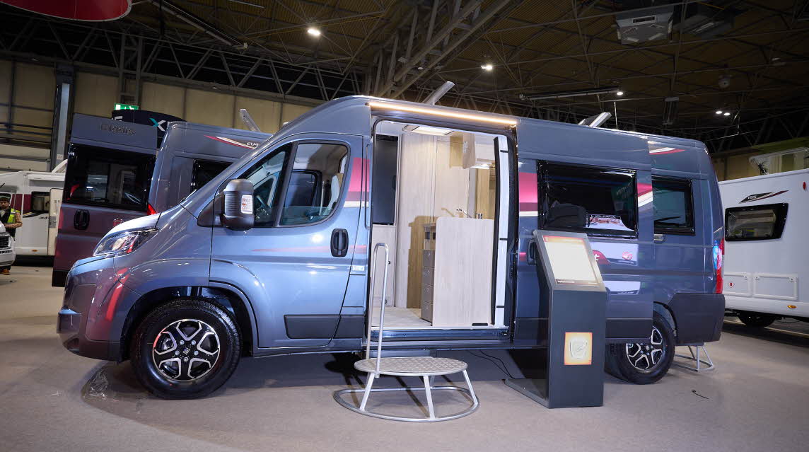 The Elddis Autoquest CV20 is blue.  There is a metal step in front of the sliding habitation door which is open revealing the interior.  There is an interactive stand to the right.
