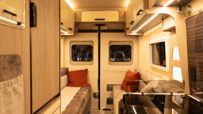 The WildAx Motorhomes Meteor has cream and brown upholstery with cream glossy cabinets.  The hob and oven are to the bottom right with the high level fridge opposite.  There is an orange cushion on each sofa.