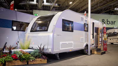 The Adria Altea 622 DK Avon’s exterior has light grey walls with a patterned decal from the front side window to the rear.  There are two wooden steps with an interactive display to the right. There is a floral display around its hitch