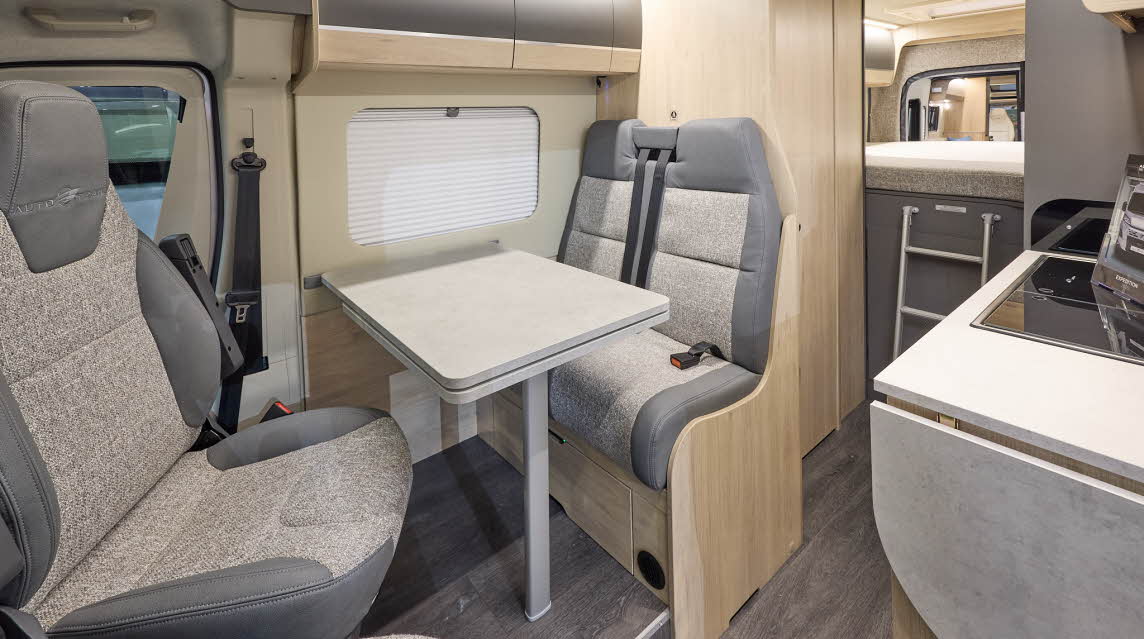 The Auto-Trail Expedition 68 has grey flooring, with pale wood cabinets with pale wood and grey overhead lockers.  The upholstery is grey and cream.  There is a ladder leading to the rear fixed bed.