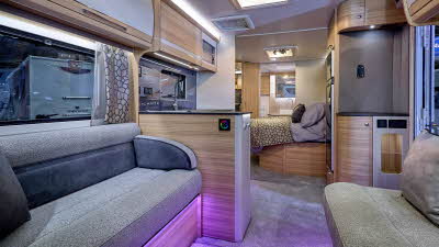 Bailey Alicanto Evora’s interior, it is grey with light wood.  The upholstery is grey and there is purple accent lighting.  The fixed island bed is beyond the kitchen and the washroom door at the rear is open.