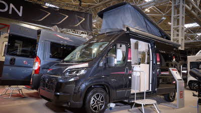 The Elddis Autoquest CV80 is black.  There is a metal step in front of the sliding habitation door which is open revealing the interior.  There is an interactive stand to the right.  The pop top roof is fully extended.