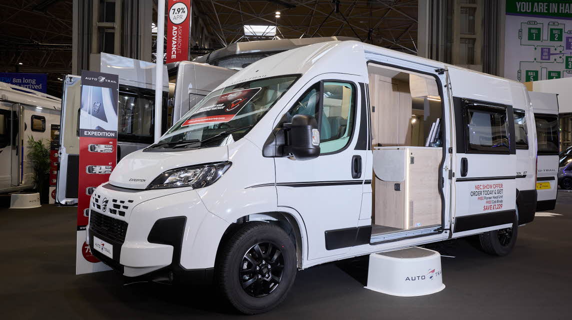 The Auto-Trail Expedition 67 is white with black decals.  There is a white step by the open sliding door which reveals the interior.  Its rear doors are open.