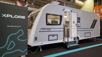 The Xplore XC QUAD is white with grey sides and black decals.  There are two metal steps to gain easy access.  There is a large Xplore advertising banner next to the QUAD. 