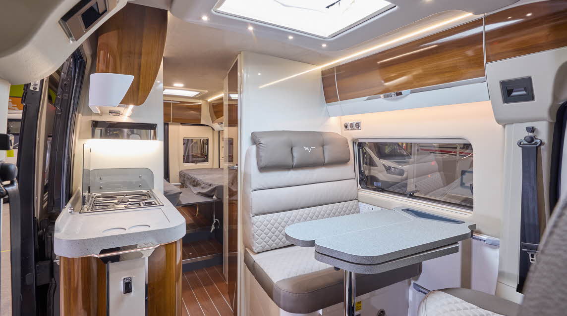 The Mobilvetta Admiral K6.3 has wooden flooring with high gloss wooden cabinets.  The upholstery is white and beige.  There is a grey table in front of the sofas.  There are two steps to the fixed rear beds.  
