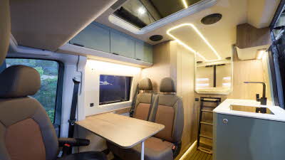 The Kaktus Adventure Vans Dune x4 has a wooden floor. The upholstery is two tone brown leather. The cabinets are grey. There is a ladder to the fixed rear bed. The pop top roof is extended.