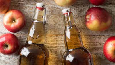 Two see through glass bottles filled with cider, laying on the table with two red apples laying by the outside of each bottle