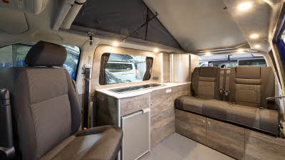 The Rolling Homes Camper Ltd Shackleton has beige flooring.  The upholstery is brown with a matching pattern.  The cabinets are wooden.  The rising roof is fully extended.