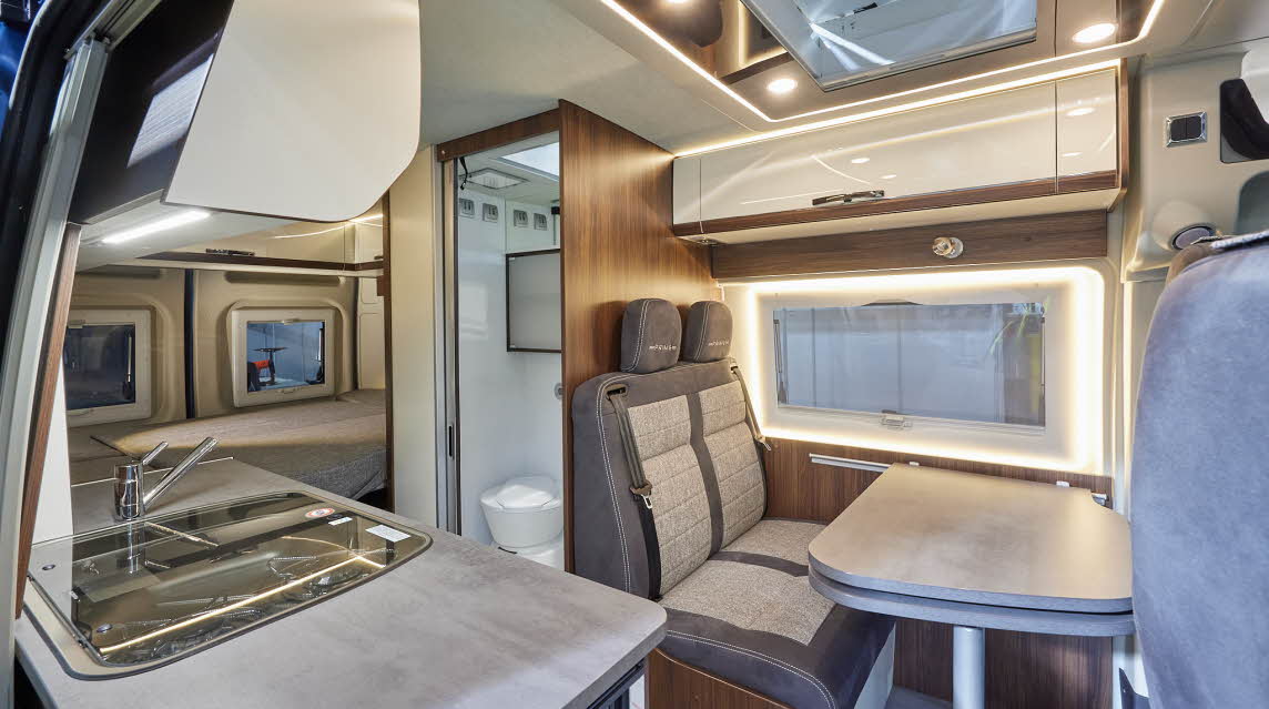 Globecar Summit Prime 600 has two tone grey and beige upholstery, cream and wooden overhead lockers.  The kitchen work surface is in grey with the central washroom opposite.  There is a fixed bed at the rear.