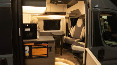 The VANTourer 600 D has brown and cream upholstery with matching cabinets.  The kitchen unit is in front of the sliding door and the lid to the hob is up.