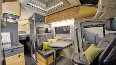 Adria Twin Max's interior has brown upholstery with curved wooden units.  There are four lime green cushions, it has a clear sunroof and the driver’s and front passenger's seat are swivelled.  The fixed bed is to the rear with the hob on the left.