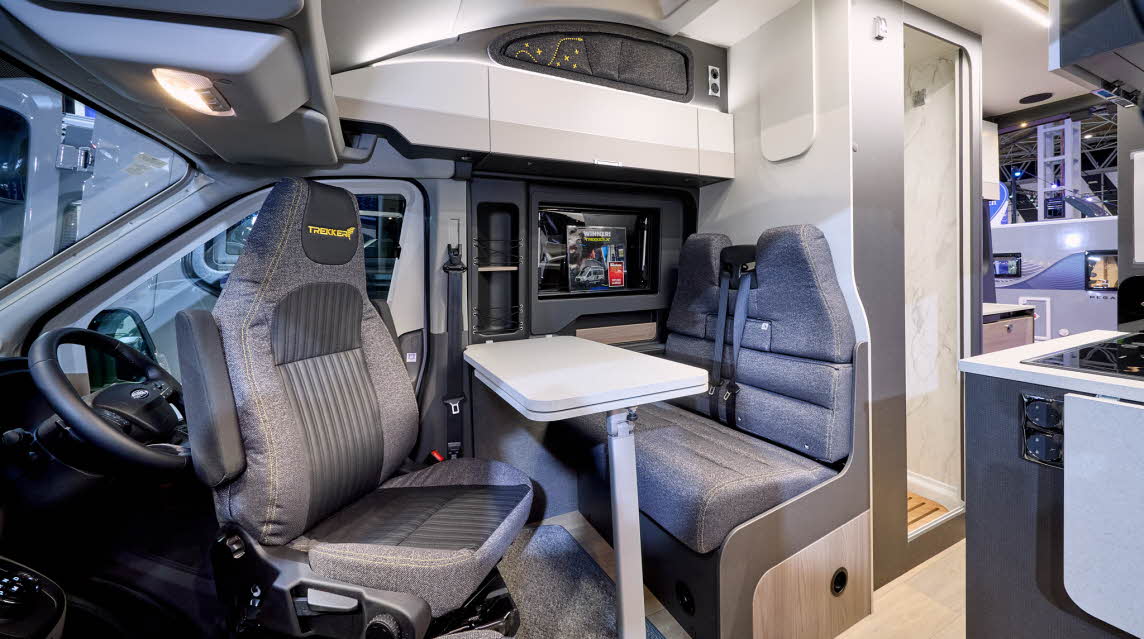 The Swift Trekker X has wooden flooring with grey upholstery.  The cabinets are white.  The door to the washroom is open.  The rear doors are open too.