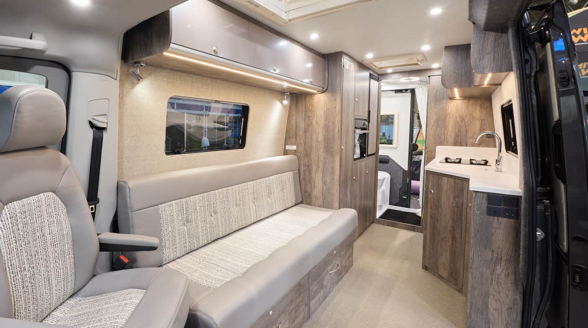 The Rolling Homes Camper Ltd Darwin FL has beige carpet.  The upholstery is mid brown with a stripped pattern.  The cabinets are pale brown with wooden surrounds.  The interior door is open revealing the rear washroom.  There are two roof lights.
