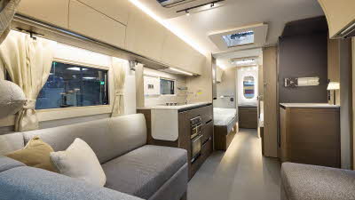 Interior of the Adria Alpina Colorado with grey sofas, cosy cushions and wood accents, illuminated by natural light through windows and skylights, offering a view outside.  Perfect for relaxation and designed for comfort.