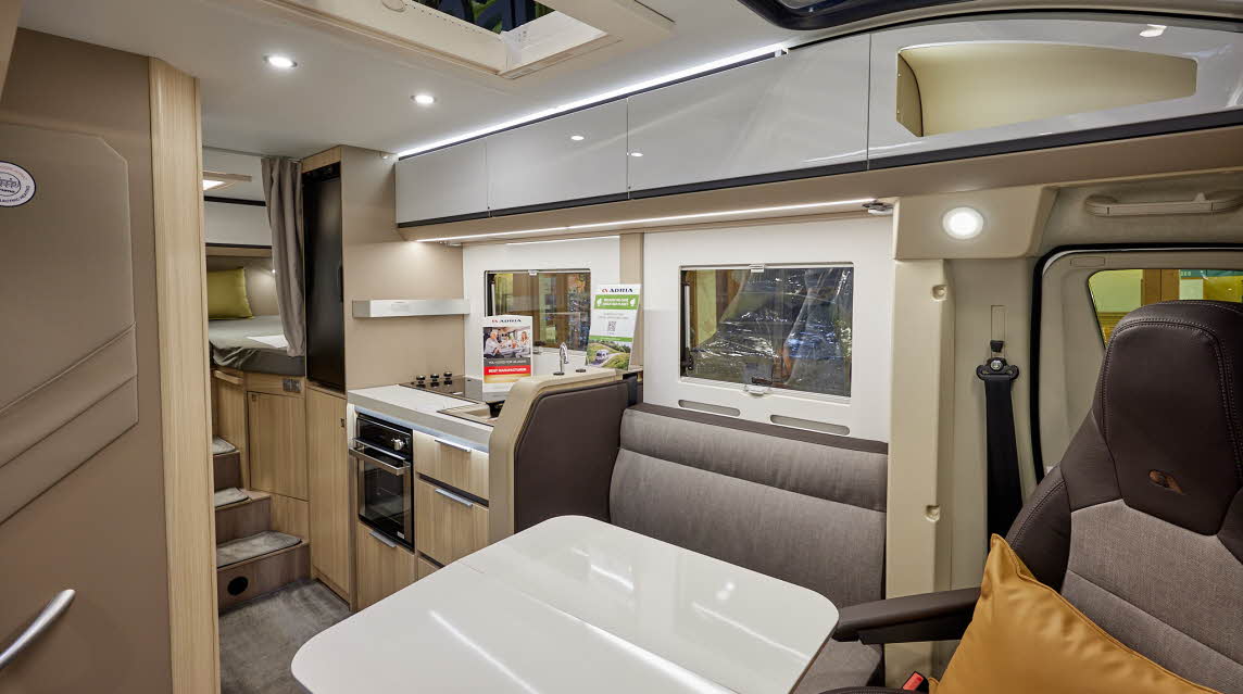 Adria Compact Supreme DL has grey upholstery, with shiny white overhead cabinets and table.  It has light wooden panelling, there are mid-brown carpets, there are three steps leading to the fixed rear bed with a privacy curtain.