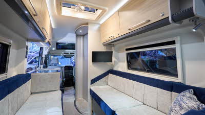 Bailey Endeavour B62’s interior has beige and dark blue fabric upholstery with blue/white cushions.  There is a TV above the left hand sofa.  The furniture is wooden.  There’s one sky light in the fixed roof.  