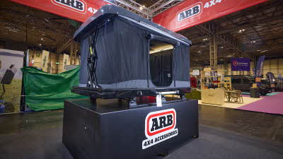 The ARB 4x4 Accessories Altitude is fully extended with two crossed mechanical poles. It has a solid grey roof and the sides are made of grey canvas. The door is open, revealing a pocket and a closed window. The unit is on car roof bars.