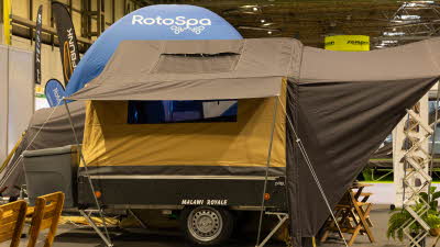 The Cabanon Malawi 2.0 Royale has brown canvas with yellow panels.  Its awning has chairs, a table, an information stand and plants.  The rear has a storage box.