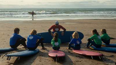 Six children sitting on their surfboards on the sand facing the surfing instructor, with a surfer walking into the sea waves in the background
