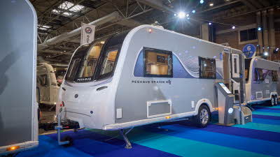 Bailey Pegasus Grande GT75 Ancona’s exterior has grey sides with a blue and white patterned decal from the front to the rear.  The entrance door is open and there are two metal steps to gain easy access.  There is an interactive display board.