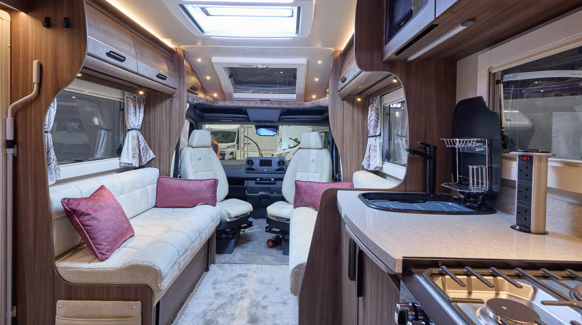 The Auto-Sleeper Burford has a pale grey carpet, with dark wood cabinets.  There are three burgundy cushions.  There is a gas hob with a tower of sockets nearby.  The driver and passenger seats are swivelled and face the interior.