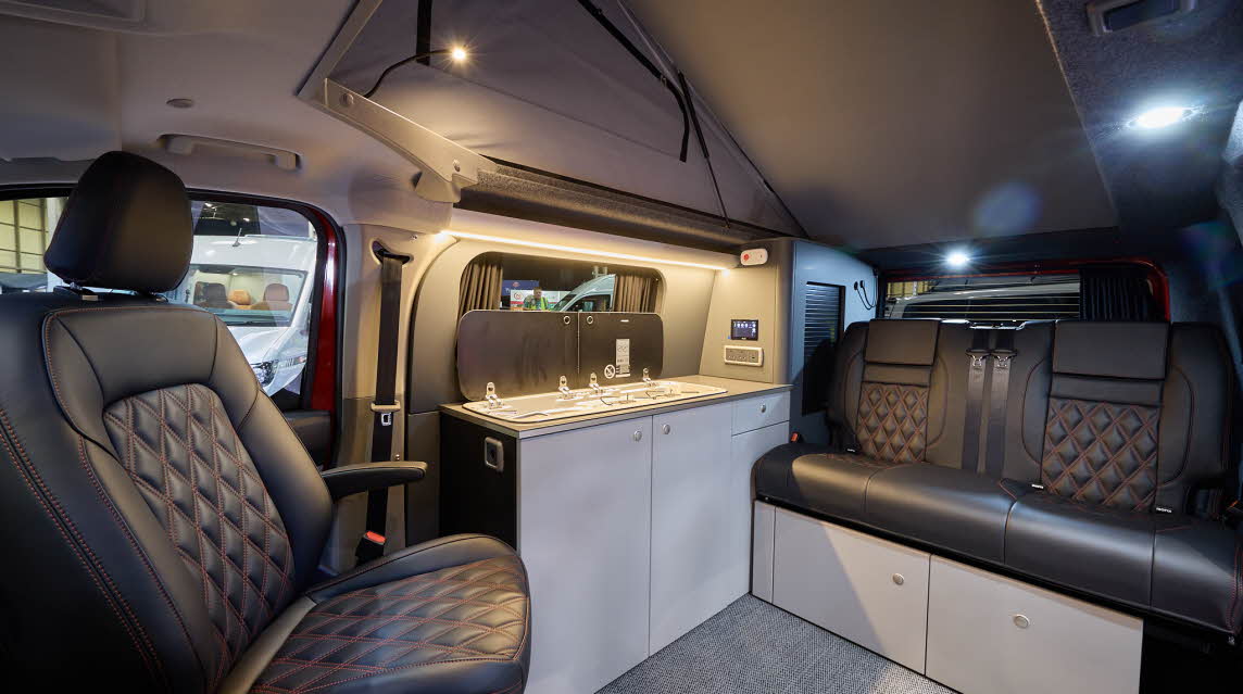The Bespoke Campers Explore has a grey carpet, with pale grey cabinets.  The upholstery is black leather with red stitching.  Its pop top roof is fully extended. 