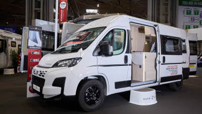 The Auto-Trail Expedition 67 is white with black decals.  There is a white step by the open sliding door which reveals the interior.  Its rear doors are open.