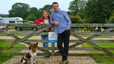 Man in blue shirt standing next to woman in red jacket in front of gate with their dog nearby