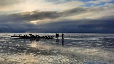 Reflective shallow sea with a large piece of driftwood and 3 people next to it like a silhouette in a darkening summer sky 