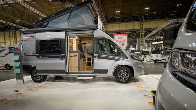 Malibu Van diversity GT skyview 600’s exterior is silver with back and gold decals.  Its sliding door is open revealing the lounge and silver ladder.  There is a step to gain easy access.  The rising roof is fully open showing a grey canvas lining.