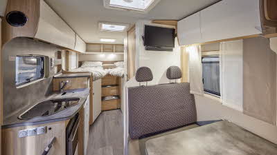 Frankia MT7 GDK Neo has patterned brown upholstery, cream and wooden overhead lockers.  The kitchen work surface is in grey with a hob and separate sink.  There is a fixed bed at the rear.  There are three roof lights.