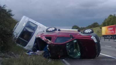Picture shows a car and a towed caravan tipped on its side by the side of a road