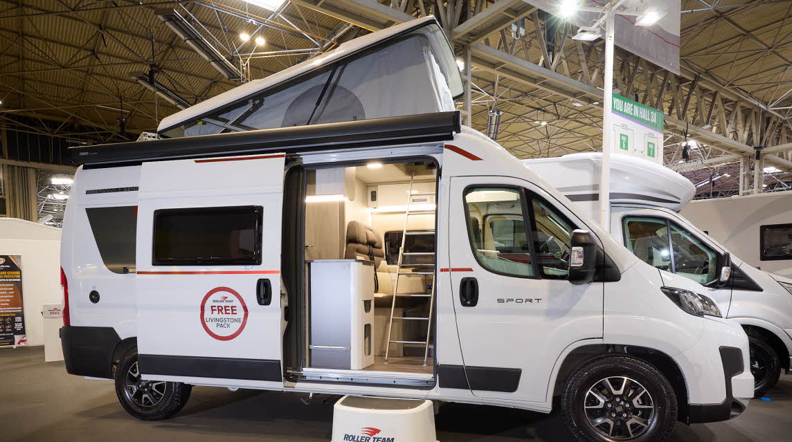 The Roller Team Livingstone 2 has a white exterior with black trim.  There is a white step outside the open sliding door revealing the interior.  The pop top roof is fully extended.