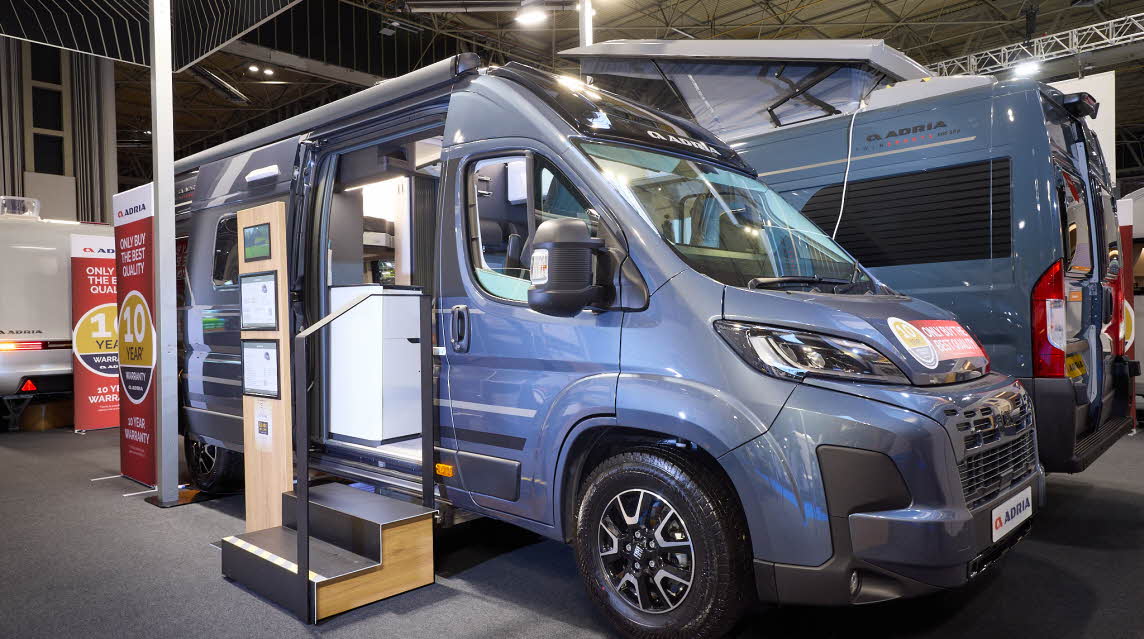 The Adria Twin Supreme has a blue exterior.  The habitation door is open and there are two steps to gain easy access. There is an information board to the left of the steps.  