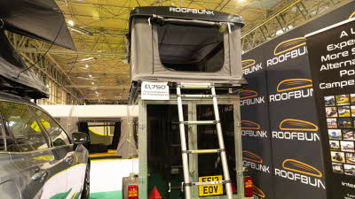 The RoofBunk Roamer has beige canvas with a black hard shell roof.  It has a silver ladder to gain access.  Its front door is open.