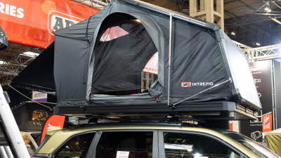 The ARB Intrepid has black canvas with a black hard shell roof.  Its door is open.  It is displayed on a gold car.