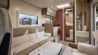 The Roller Team Kronos Compact 590 has grey carpet. The upholstery is cream and mid brown. The cabinets are cream with wooden surrounds and the interior door is a dark brown. There is a step up to the rear washroom.