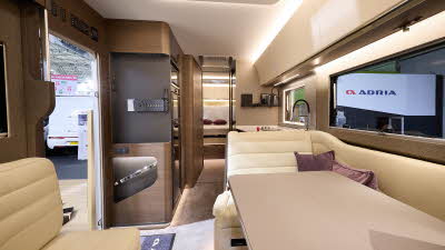 Adria Supersonic's interior has cream upholstery with curved dark wooden units.  There are two dark pink cushions, it has a sky light and a beige table.  The fixed bed is to the rear and there is a place for a TV on the left hand side.