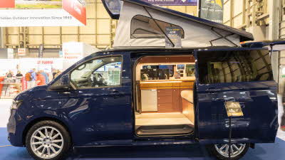 The Out and About Campers Alpine Tourer is dark blue, its rising roof and sliding door are open.  There is a brochure stand next to it.