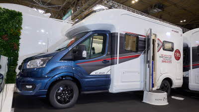 The Auto-Trail F-Line F62 has a blue cab and white body with grey and red decals.  There is a show offer on its side in red.  There is a white step by the opened habitation door.  
