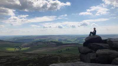 A person sitting high on a rock on the top of Stanage Edge looking out over the rolling hills that go on more miles under a blue sky of white clouds