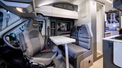 The Swift Trekker X has wooden flooring with grey upholstery.  The cabinets are white.  The door to the washroom is open.  The rear doors are open too.