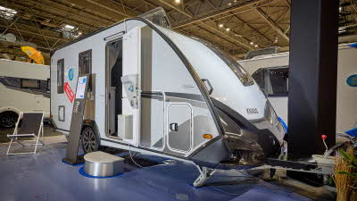 The Knaus Sport&Fun 480 QL Black selection is white with black/grey edging. The entrance door is open and there is a metal step to gain easy access. There is a stand next to the steps with a deck chair just beyond.