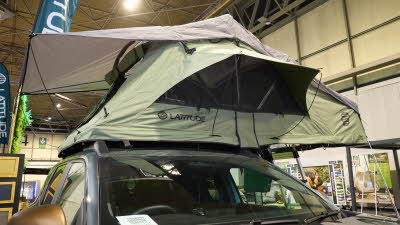 The Latitude Tents Pioneer has green canvas walls and a pale grey roof. The window is open. It has an extended platform with silver ladder to gain easy access